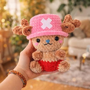 May include: A handmade plush toy of a cartoon character, featuring a pink hat with a white cross, brown antlers, and red shorts. The toy has a light brown body, black eyes, and a blue nose. The toy is approximately 6 inches tall.