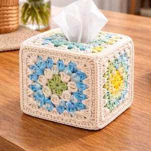 May include: A square, handmade tissue box cover. The cover is crocheted with a cream-colored yarn and features a floral pattern in shades of blue, green, and yellow. White tissues are visible at the top. The box sits on a wooden surface.