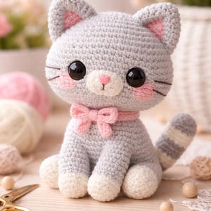 Op de afbeelding: Een handgemaakte gehaakte kattenpop in grijs met roze accenten. De kat heeft grote zwarte ogen, roze wangen en een roze strik. De poten en oren zijn ook roze. De kat zit op een houten oppervlak, met garen en knutselspullen op de achtergrond.
