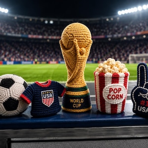 May include: A collection of crochet items related to soccer and the USA. Includes a soccer ball, a miniature USA jersey, a World Cup trophy, a popcorn container, and a foam finger with "#1 USA" text. The items are displayed on a blue surface.