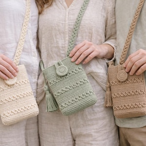May include: Three crochet crossbody phone bags in cream, sage green, and tan. Each bag has a matching strap, a decorative button closure, and a tassel. The bags are made of a textured yarn with a geometric pattern.