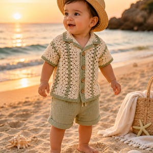 May include: A baby wearing a straw hat, a cream-colored crochet shirt with green trim, and matching green shorts stands on a sandy beach. The shirt has wooden buttons. A woven basket, seashells, and a starfish are also on the beach.