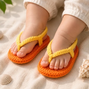 Può includere: Un paio di sandali per neonati all'uncinetto arancioni e gialli. I sandali sono sui piedi di un bambino, appoggiati su una superficie sabbiosa. I sandali hanno un design a infradito, con un cinturino giallo e una base arancione.