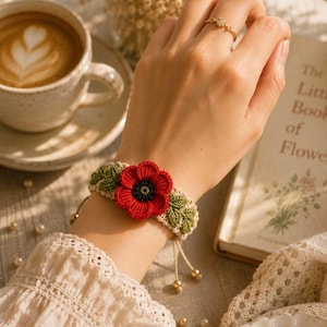 K&ouml;nnte beinhalten: Handgefertigtes H&auml;kelarmband mit einer leuchtend roten Mohnblume und gr&uuml;nen Bl&auml;ttern. Das Armband ist beige mit goldfarbenen Perlen und einem Kordelverschluss. Ein goldener Ring mit floralem Design ist ebenfalls zu sehen.
