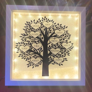 May include: A framed family tree artwork with a black tree silhouette. Heart-shaped leaves bear names such as Grace, Max, and Oliver. The background is glittery gold, and the white frame has embedded lights. A decorative piece.