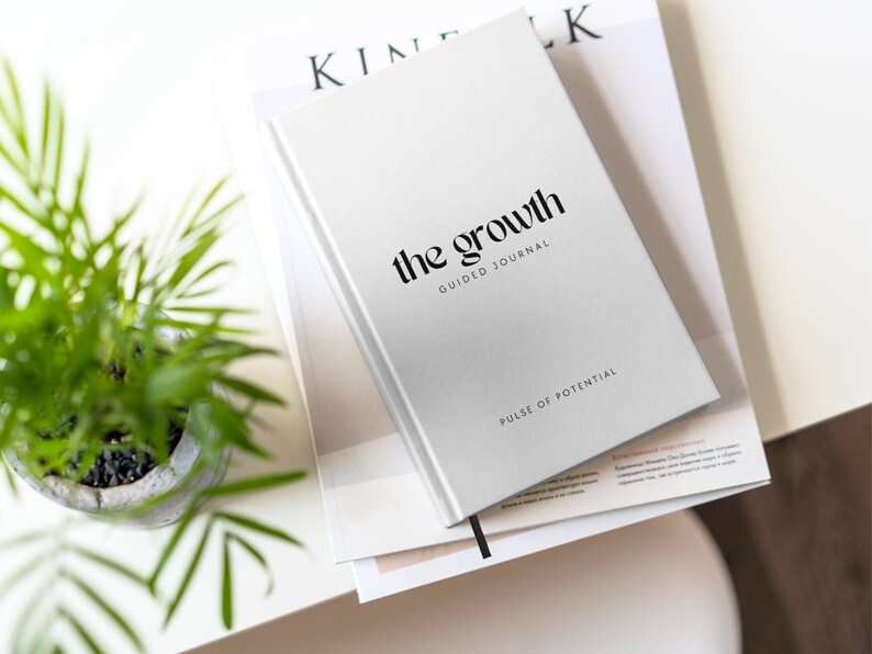 May include: A white journal with the text "the growth guided journal" and "pulse of potential" on the cover. The journal is stacked on top of a magazine.