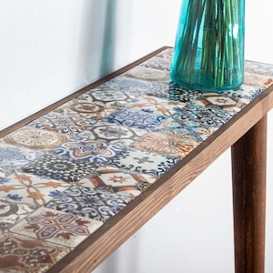 Può includere: Un tavolino da console in legno con un piano in piastrelle. Le piastrelle sono un mix di blu, bianco e marrone con motivi intricati. Il tavolo ha un design semplice con due gambe.