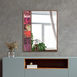 May include: A rectangular wall mirror with a wooden frame. The left side of the frame features a collage of colorful floral images. The mirror reflects a window with a sheer curtain and indoor plants. The mirror is mounted on a gray wall.