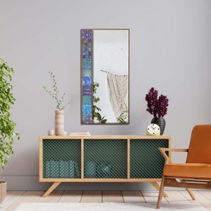 May include: A rectangular mirror with a brown frame and a decorative mosaic tile border in shades of blue, purple, and orange. The mirror reflects a room with a white wall, a macrame hanging, and a wooden cabinet.