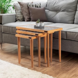 May include: A set of three wooden nesting tables with square tops. The tables have light brown legs and patterned tops. A gray sofa and a potted plant are in the background.