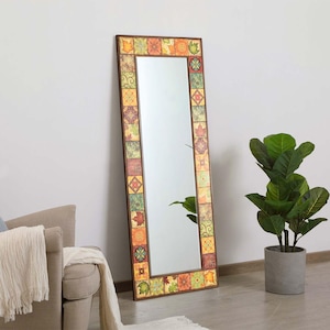 Op de afbeelding: Een rechthoekige spiegel over de volledige lengte met een decoratief frame. Het frame bestaat uit kleine, kleurrijke vierkante tegels in tinten oranje, groen en bruin. De spiegel leunt tegen een witte muur, naast een potplant.