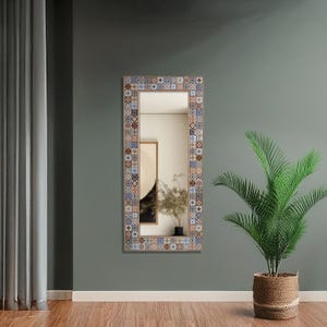 Può includere: Uno specchio da parete rettangolare con un bordo decorativo con un motivo ripetuto di piastrelle blu, marroni e bianche. Lo specchio è montato su una parete verde, con una pianta in vaso in primo piano.