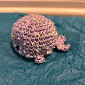 May include: A small crocheted whale toy with a purple body and colorful speckles. The whale is lying on a blue textured surface.