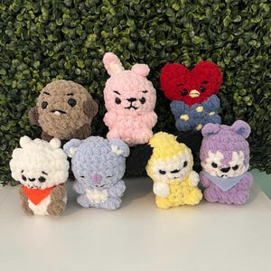 May include: A collection of handmade crochet plushies in various colors and designs. The plushies include a brown dog, a pink bunny, a red heart, a white bear, a blue koala, a yellow duck, and a purple wolf. They are arranged in a group.
