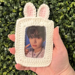 May include: A white, crocheted photo frame with bunny ears. The frame holds a photo of a person. The bunny ears are white with pink inner ears. The frame is held in a hand against a background of green foliage.