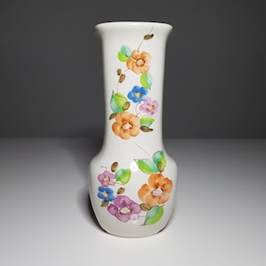 Può includere: Un vaso in ceramica bianca con un motivo floreale. Il vaso ha un collo stretto e una base larga. I fiori sono dipinti in tonalità di arancione, rosa, blu e verde.
