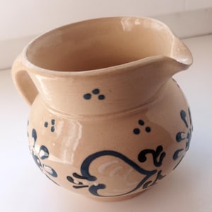 Op de afbeelding: Een beige keramische kan met een handvat en een tuit. De kan is versierd met donkerblauwe bloemen- en stippenpatronen. De kan heeft een afgeronde body en een licht uitlopende rand.
