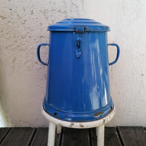 Può includere: Un contenitore vintage in smalto blu con coperchio, due maniglie laterali e un fermo. Il contenitore è cilindrico con una base leggermente svasata e poggia su uno sgabello bianco. Lo smalto presenta qualche segno di usura.