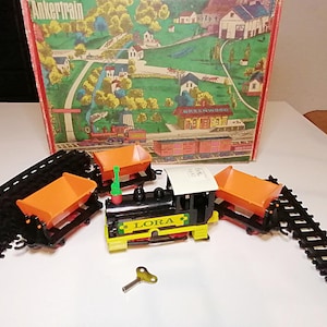 May include: A vintage toy train set with a black and yellow LORA locomotive, orange cargo cars, and black tracks. The set includes a wind-up key and is set against a backdrop of a colorful box featuring a countryside scene.
