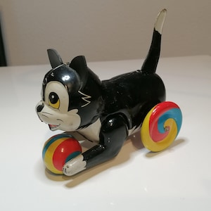 Può includere: Giocattolo vintage a forma di gatto nero con viso, orecchie e zampe bianche. Il gatto ha una lunga coda e tiene in mano una palla colorata. Le ruote hanno un design a spirale. Un giocattolo classico.