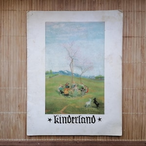 May include: Vintage print featuring a pastoral scene with children dancing around a tree on a green hill. Two small animals are in the foreground. The word "kinderland" is printed at the bottom in a decorative font.