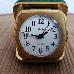 May include: A vintage gold-toned Europa travel alarm clock with a white face and black numerals. The clock has a green leatherette top and a square shape. The clock face displays the text "EUROPA" and "2 JEWELS".