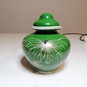 Può includere: Un vaso di ceramica verde con coperchio, con un design floreale bianco e oro. Il vaso ha una forma arrotondata e una finitura lucida. Il coperchio è anch'esso verde con un accento bianco. Un oggetto decorativo.