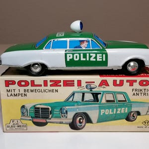 Pode incluir: Carro de polícia de brinquedo vintage em verde e branco, com a palavra "POLIZEI" na lateral. O carro está em uma caixa com as palavras "POLIZEI AUTO" e uma imagem de um carro semelhante. A caixa também tem texto em alemão.