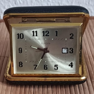 May include: A vintage Europa travel alarm clock. The clock has a silver face with black numerals and hands, a date display, and the word "EUROPA" printed on the face. The clock is encased in a gold-toned metal frame with a black leatherette cover.
