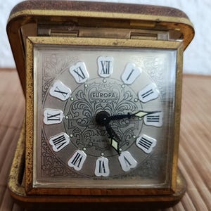 May include: A vintage Europa travel clock in a gold-tone case. The clock face is silver with black hands and Roman numerals. The clock reads "2 JEWEL" and "GERMANY". The clock is open, revealing the clock face.