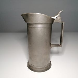 Könnte beinhalten: Ein Zinnhumpen mit Scharnierdeckel und Henkel. Der zylindrische Körper hat einen leicht ausgestellten Boden und einen Ausguss. Das Metall hat eine matte Oberfläche. Der Humpen wird wahrscheinlich zum Servieren von Getränken verwendet.