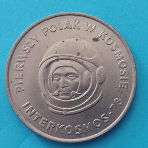 May include: A circular silver-toned commemorative coin featuring a relief portrait of an astronaut wearing a helmet. The text PIERWSZY POLAK W KOSMOSIE and INTERKOSMOS-78 is embossed along the outer edge of the coin, surrounding the central figure.