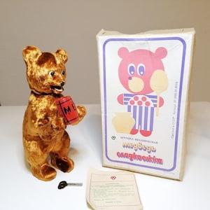 May include: A vintage, golden-brown plush bear toy holding a red barrel with the letters "ME". The bear stands next to its original box, which features a cartoon bear illustration. A key and paperwork are also present.