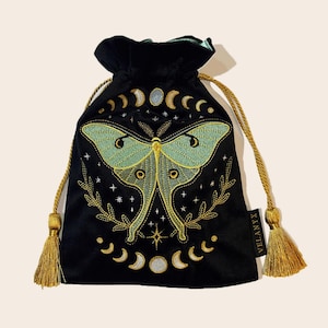 May include: A black velvet drawstring pouch featuring an embroidered mint green moth with gold accents. The design includes celestial moon phases, stars, and a laurel wreath. A black tag with the white text VELANYX is attached to the side.