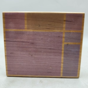 May include: A wooden block with a geometric design. The block features a combination of light brown and lavender-colored wood, with thin gold-colored lines outlining the geometric shapes. The wood grain is visible.