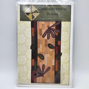 Könnte beinhalten: Ein verpacktes Quiltmuster mit dem Titel "Coneflowers in Bloom". Das Muster zeigt ein Blumendesign mit braunen und beigen Stoffen. Der Text "Featuring Jelly Roll Strips and Raw Edge Applique" ist ebenfalls sichtbar.