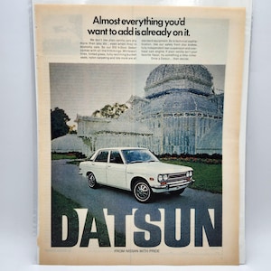 Puede incluir: Anuncio publicitario vintage de un Datsun 510 de 4 puertas blanco. El anuncio muestra el coche aparcado en una carretera con un edificio al fondo. La palabra "DATSUN" se muestra de forma destacada en letras grandes, con el texto "FROM NISSAN WITH PRIDE" debajo.