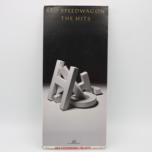 May include: A compact disc case for REO Speedwagon's album "The Hits." The case is primarily black and white, with the band's name and album title in gold lettering. The word "HITS" is displayed in large, 3D white letters.