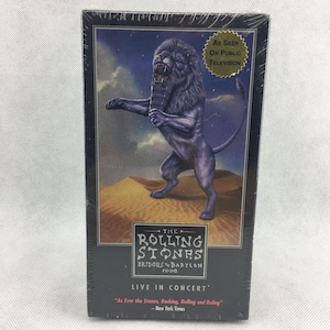 Può includere: Videocassetta VHS sigillata del concerto "Bridges to Babylon" dei Rolling Stones del 1998. La copertina presenta un'illustrazione di un leone viola con la scritta "As Seen On Public Television". Il nome della band e il titolo dell'album sono ben visibili.