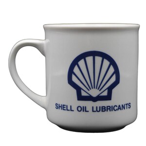 May include: White ceramic mug featuring the Shell Oil logo in blue, a stylized scallop shell design. The words "SHELL OIL LUBRICANTS" are printed below the logo. The mug has a curved handle.