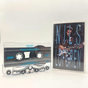May include: A clear cassette tape with a black interior and white spools is next to a cassette case. The case features a photo of a man playing a guitar, with the name "NILS LOFGREN" in blue text.