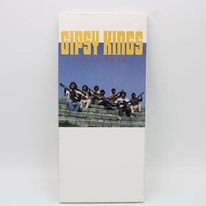 May include: Album cover for "Gipsy Kings Allegria" featuring a group of musicians with guitars. The top portion has the band's name in yellow and blue over a blue sky background. The musicians are seated on stone steps.