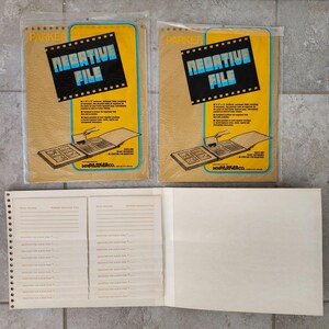 May include: Two yellow and brown paper envelopes with the text "NEGATIVE FILE" printed on them. The envelopes are for storing photographic negatives. The envelopes are open and show a brown photo album with a white page inside. The album has a white spine with the text "BENJA PARKER CO." printed on it. The album is open to a page with a grid of squares for storing negatives. The album is on a white surface.