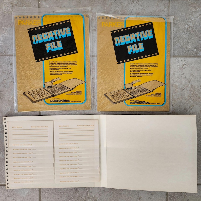 May include: Two yellow and brown paper envelopes with the text "NEGATIVE FILE" printed on them. The envelopes are for storing photographic negatives. The envelopes are open and show a brown photo album with a white page inside. The album has a white spine with the text "BENJA PARKER CO." printed on it. The album is open to a page with a grid of squares for storing negatives. The album is on a white surface.