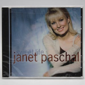 May include: CD case featuring Janet Paschal with the album title "Sweet Life." The cover art showcases a smiling woman with blonde hair against a blue and white background. The artist's name is in a copper color.