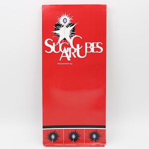 May include: A tall, rectangular box with a vibrant red background. The box features the text "SUGAR CUBES" in large, white, stylized letters, with the tagline "Stick Around For Joy" below. The top has a graphic of a starburst with a blue sphere.