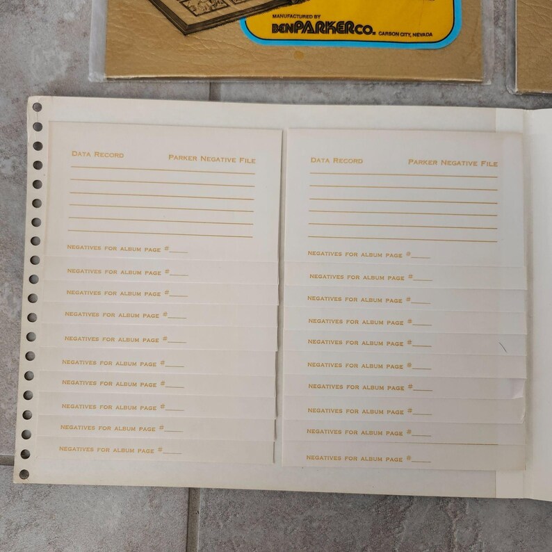 May include: A white photo album with a beige cover and gold lettering. The album has a data record section and a section for listing negatives for album pages. The album is manufactured by Ben Parker Co.