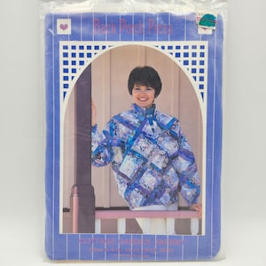 以下が含まれることがあります： 青、紫、白のキルティングジャケットを着た女性が写っている、縫製パターンのパッケージ。パッケージには「Back Porch Press」と「Cottage Garden Jacket」の文字が入った青い縁取りがあります。ジャケットはハイネックです。