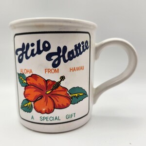 May include: White ceramic mug with the words "Hilo Hattie" in blue script, "Aloha From Hawaii," and a vibrant orange hibiscus flower. The mug has a curved handle and the text "A Special Gift."