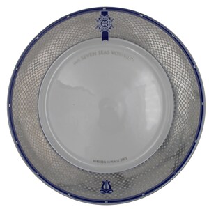 May include: A white ceramic plate with a silver and blue border. The plate has the text "The Seven Seas Voyager" and "Maiden Voyage 2003" printed on it.
